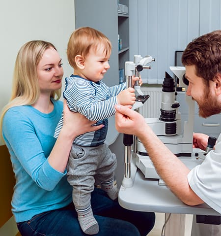 A mother holding her young infant son up to get his eyes examined by a male optometrist.
