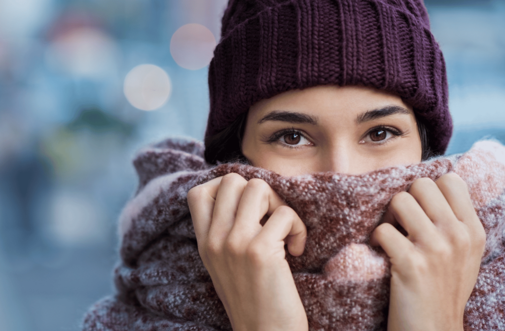 A woman wearing a knit hat, winter jacket, and a scarf, and she is holding the scarf over half of her face due to cold weather