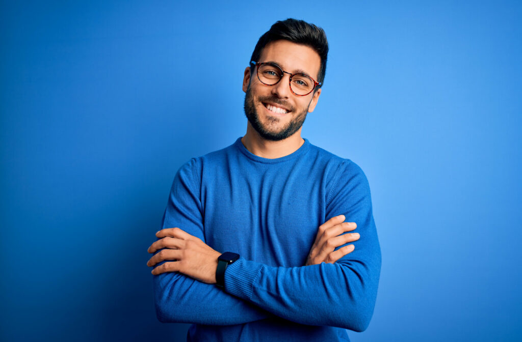 A young man wearing glasses, with his arms crossed, standing against a blue background that matches his sweater