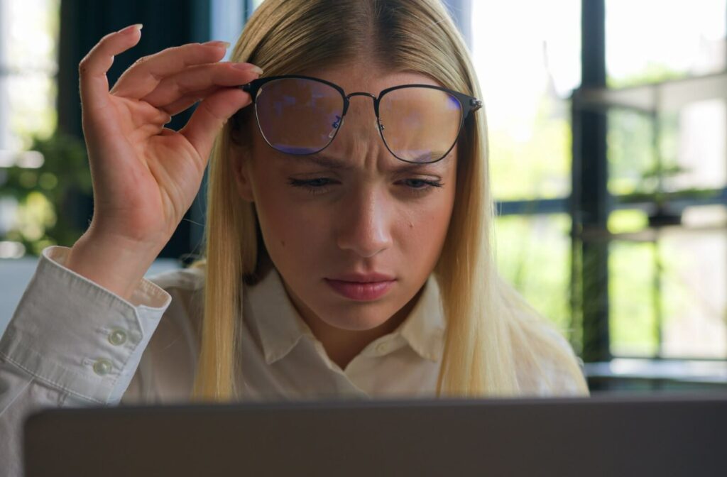 Person sitting indoors in front of a laptop, lifting their glasses while squinting at the screen with a furrowed brow, appearing to struggle with blurry vision.