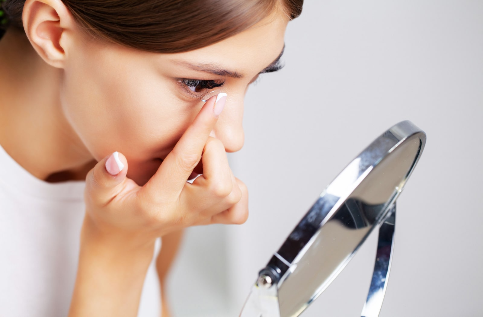 A woman looking into a mirror while putting a contact lens in.