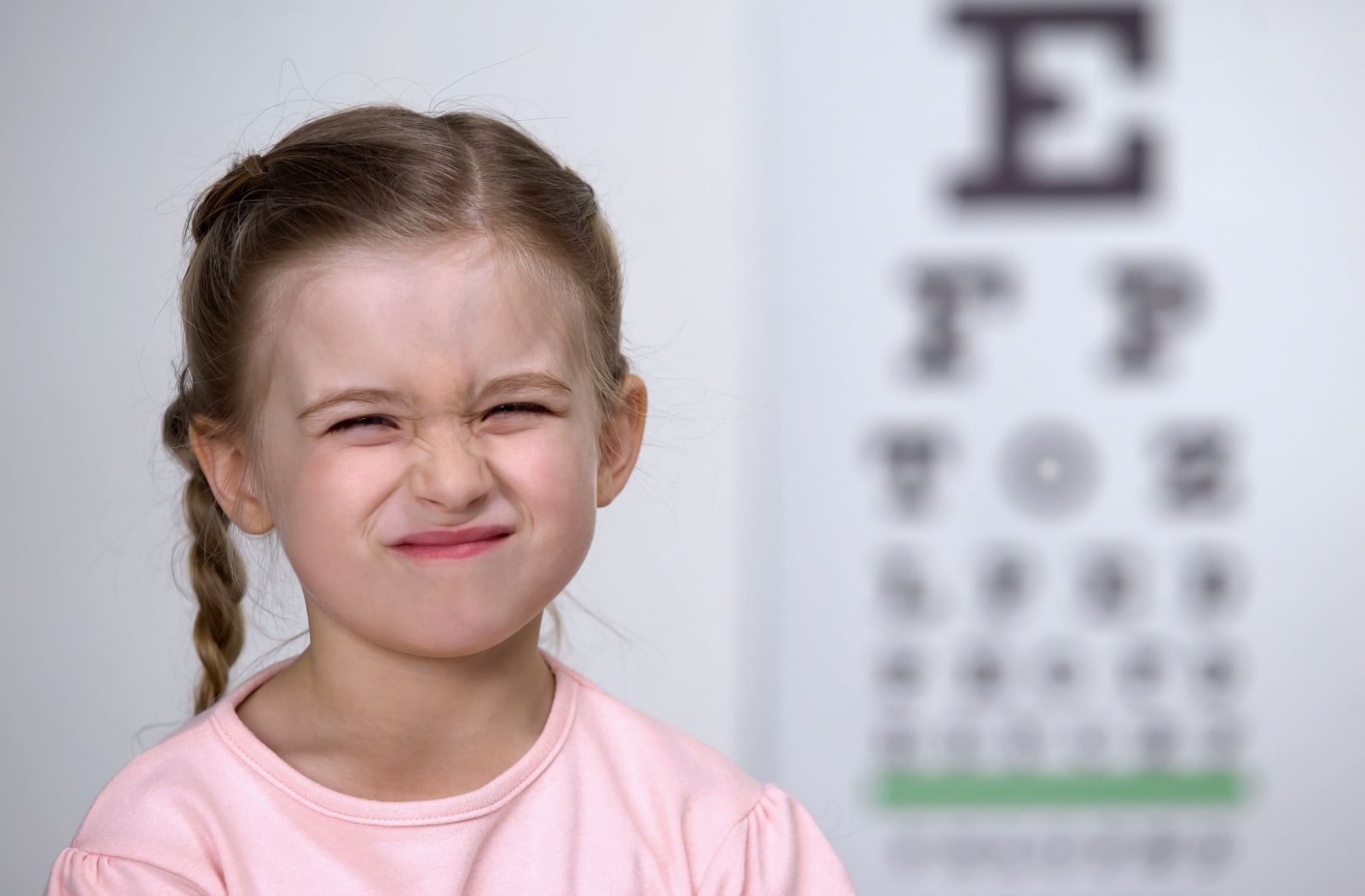 A young girl standing in front of a Snellen eye chart and squinting. Squinting may be a sign of refractive errors in your child's vision. 