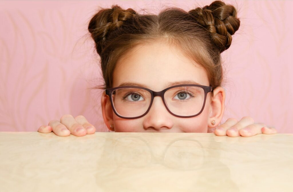 A young girl wearing corrective lenses and peaking up behind a table.