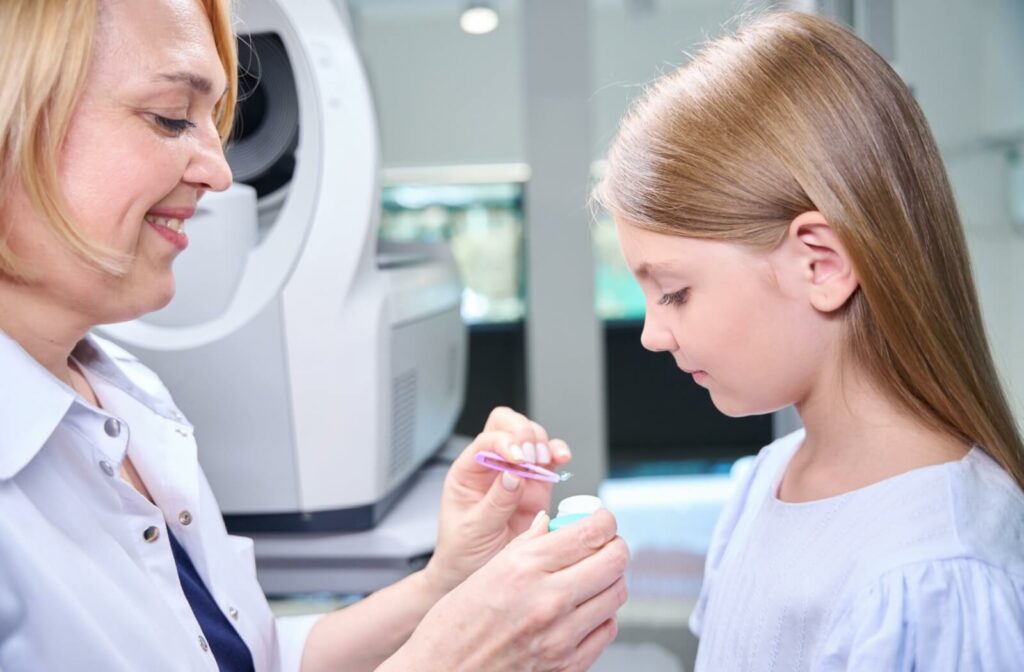 A smiling optometrist holds a contact lens with tweezers while a young child looks down at the lens.