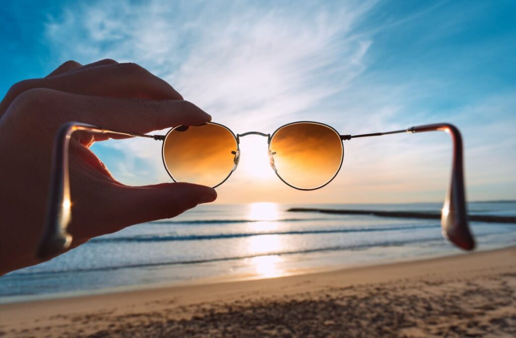 A hand holds brown polarized sunglasses up over a sunset on the beach.