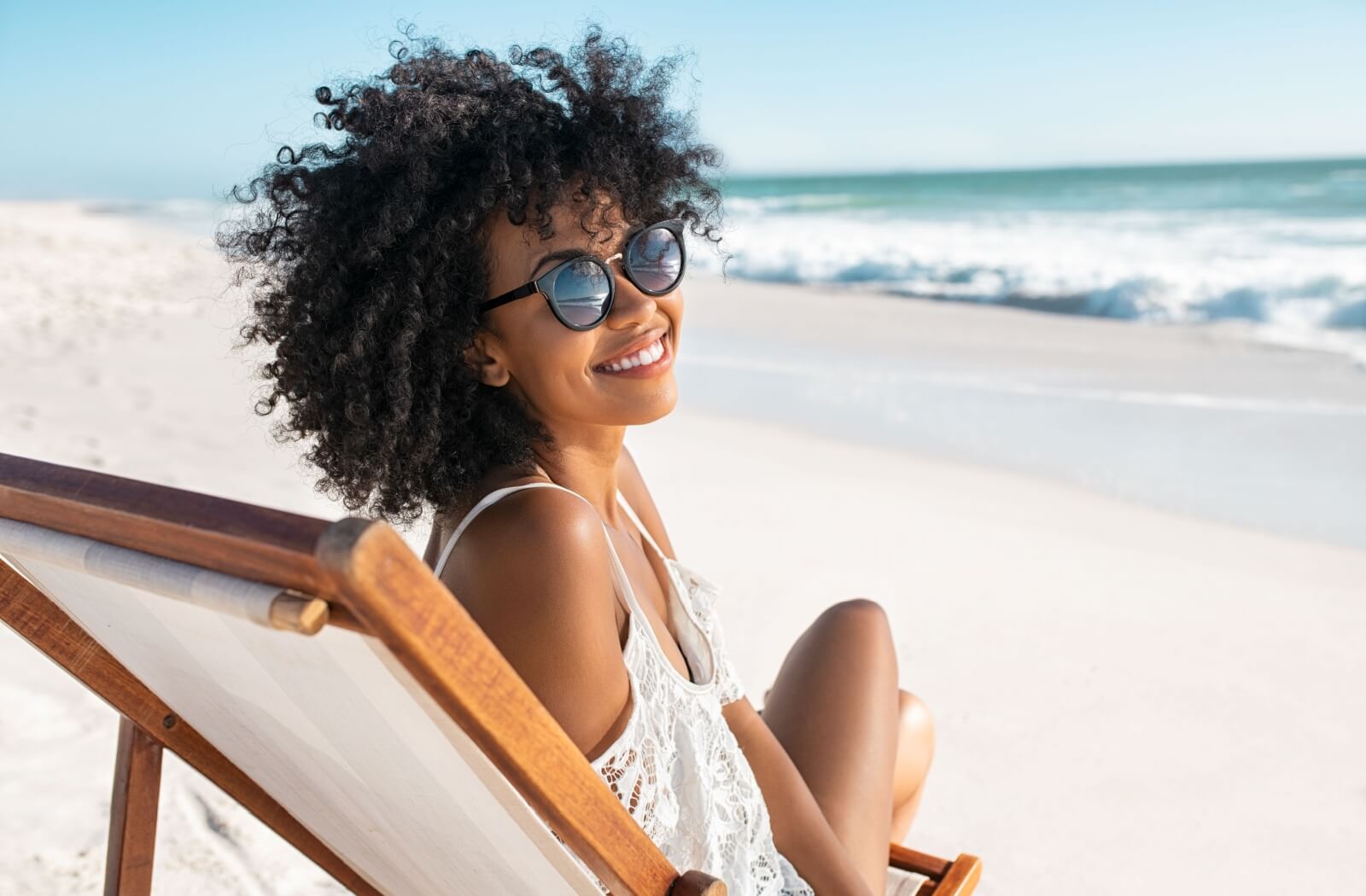 A young woman relaxes on a chair at the beach while wearing her sunglasses to protect here eyes.