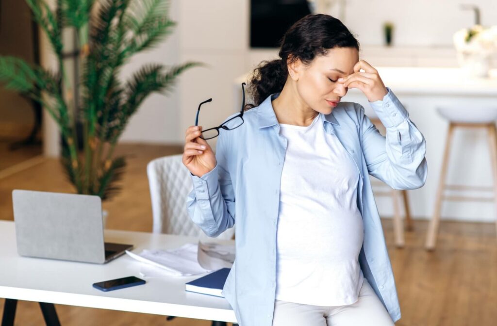 A pregnant woman at work taking off her glasses to rub her eyes in frustration due to eye twitching.