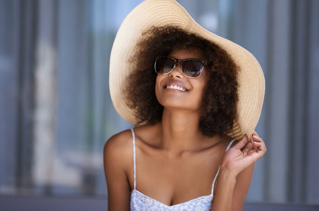 A person wearing a large, floppy hat and a pair of sunglasses smiles and enjoys a sunny day.