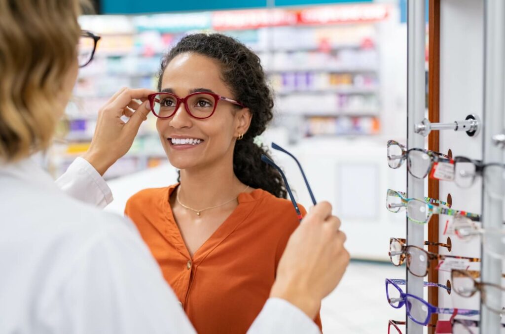 An optician assists an adult in trying on frames, offering advice on choosing between styles, colours, and frame materials.