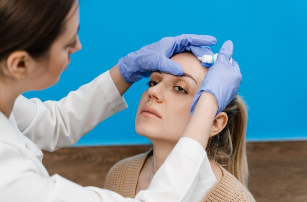 An optometrist in blue gloves applies eye drops to a patient’s eye during an examination for dry eye treatment.