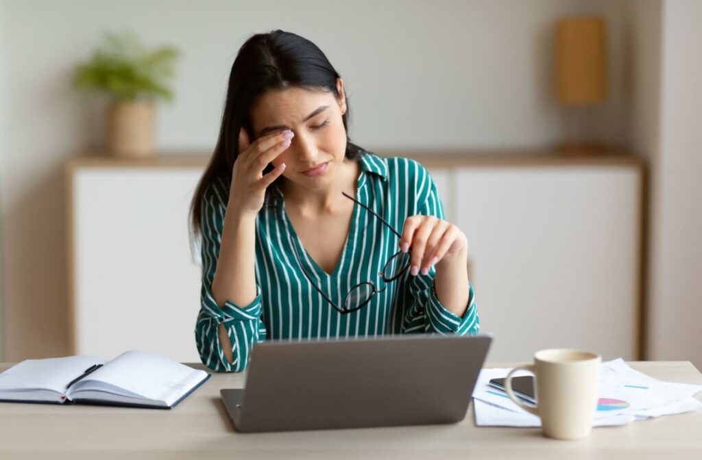 Woman in rubbing her eye while working at a laptop, experiencing symptoms of dry eye disease