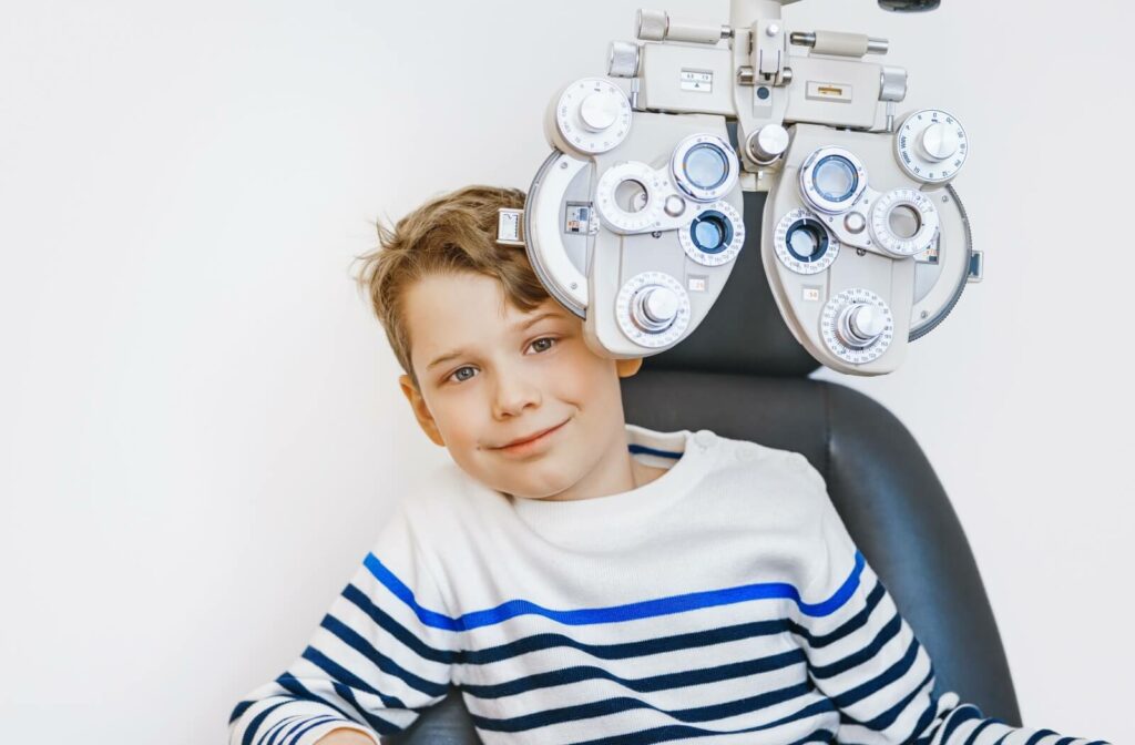 School-aged child smiling and peeking out from behind a phoropter, a large mask-like device with knobs and switches.