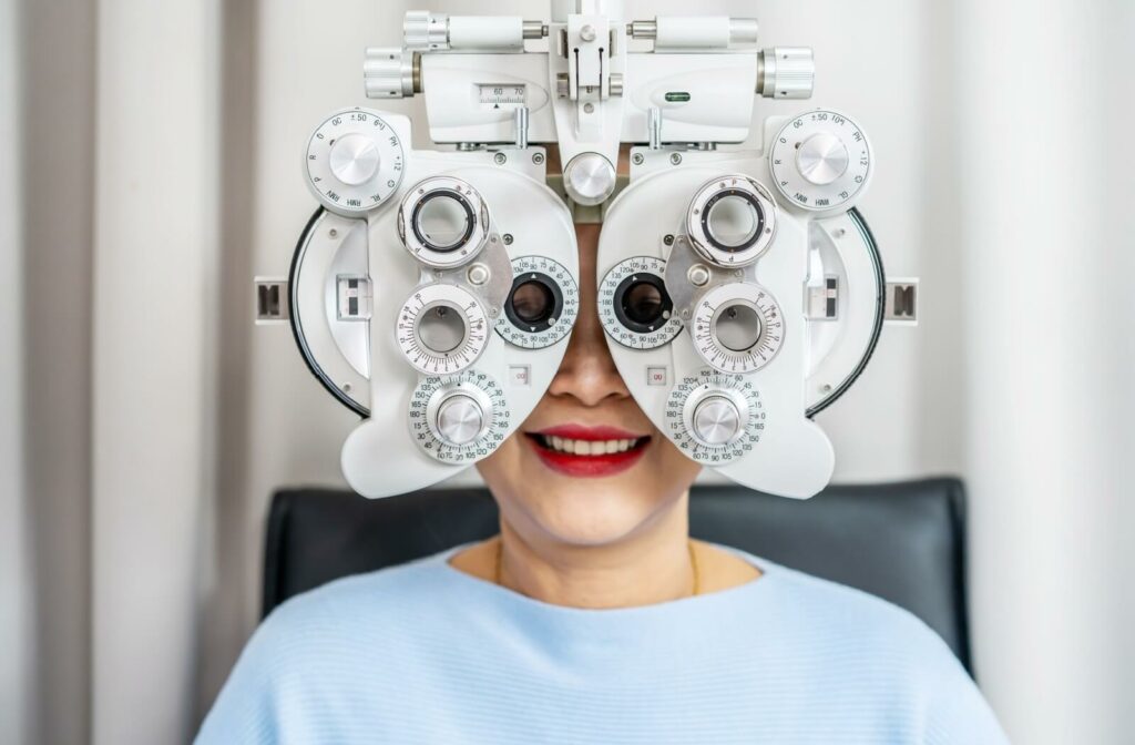 Older adult with red lipstick smiling behind a phoropter, which is an optometry device that resembles a large mask with many knobs and switches.