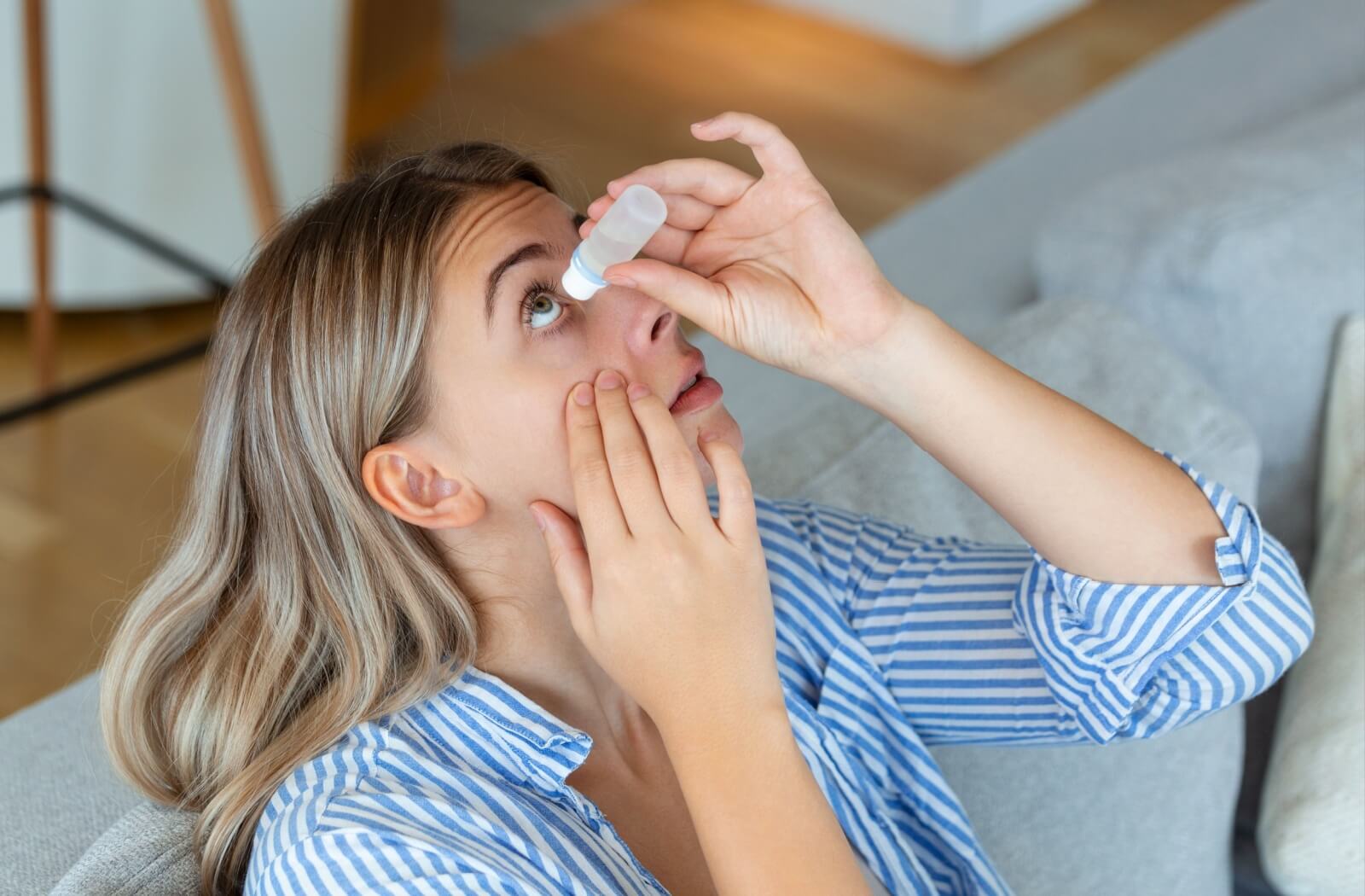 A person sitting on a couch tilting their head back while applying eye drops, holding the lower eyelid down with one hand.
