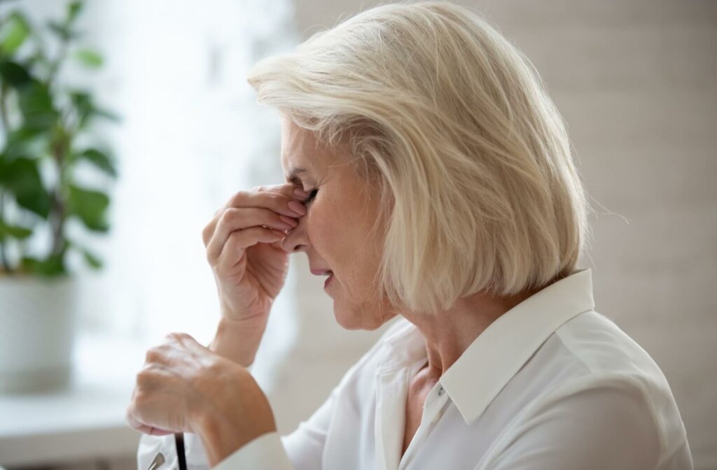 A senior with blonde hair rubbing her eyes while holding a pair of glasses, demonstrating symptoms of age-related vision loss.