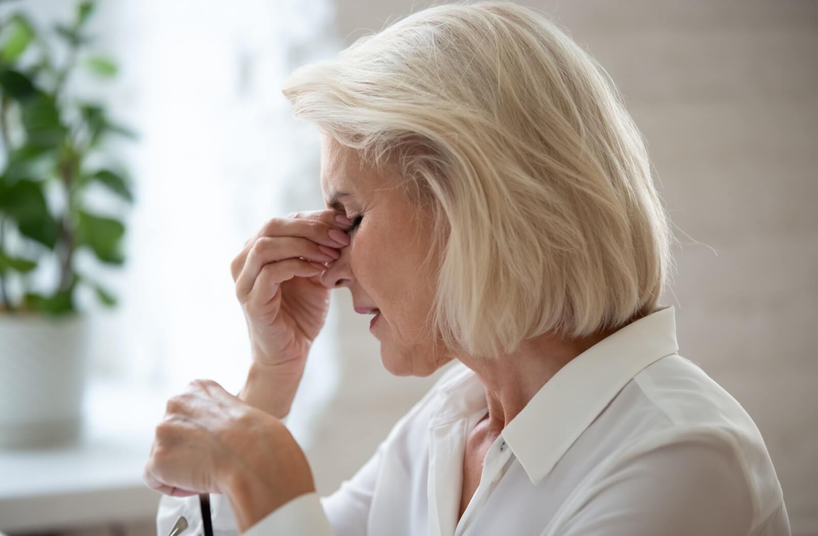 A senior with blonde hair rubbing her eyes while holding a pair of glasses, demonstrating symptoms of age-related vision loss.