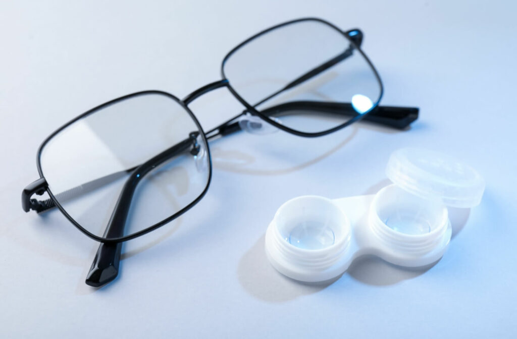A pair of black-framed eyeglasses next to an open white contact lens case containing two lenses.