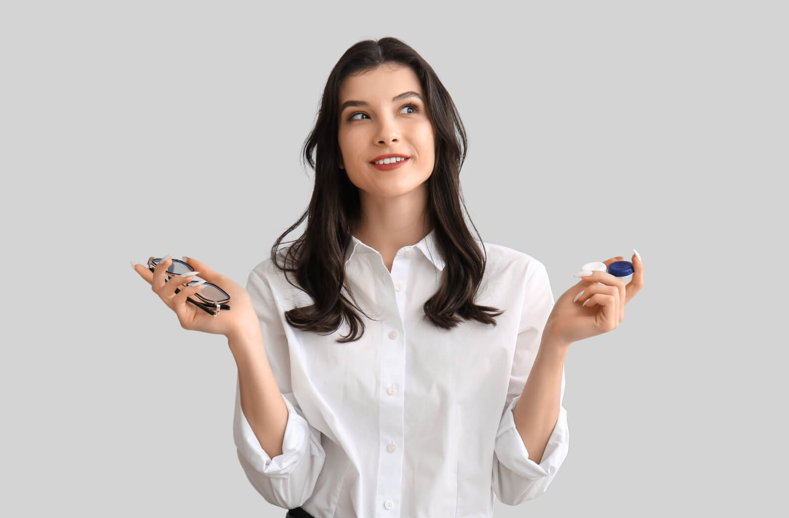 An individual holding a pair of eyeglasses in one hand and a contact lens case in the other while looking upward thoughtfully.