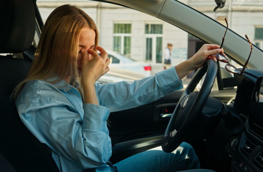 An individual sitting in the driver's seat of a car rubbing their eyes and holding their eyeglasses.