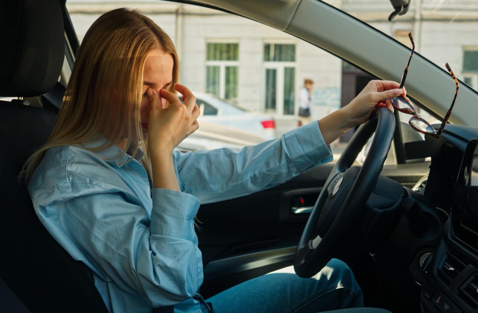 An individual sitting in the driver's seat of a car rubbing their eyes and holding their eyeglasses.